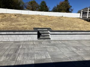 Paver patio using 24 x 24 Cambridge slabs shown in a Limestone Quarry Blend. Retaining wall was built using Cambridge Omega wall blocks shown with a Coal Platinum Smooth strip. Stairs are biult with Cambridge Crusader caps in the color Coal.