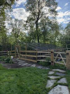 Round cedar three-rail fence with black welded wire mesh and a wooden board chicken coup with metal protection cover.