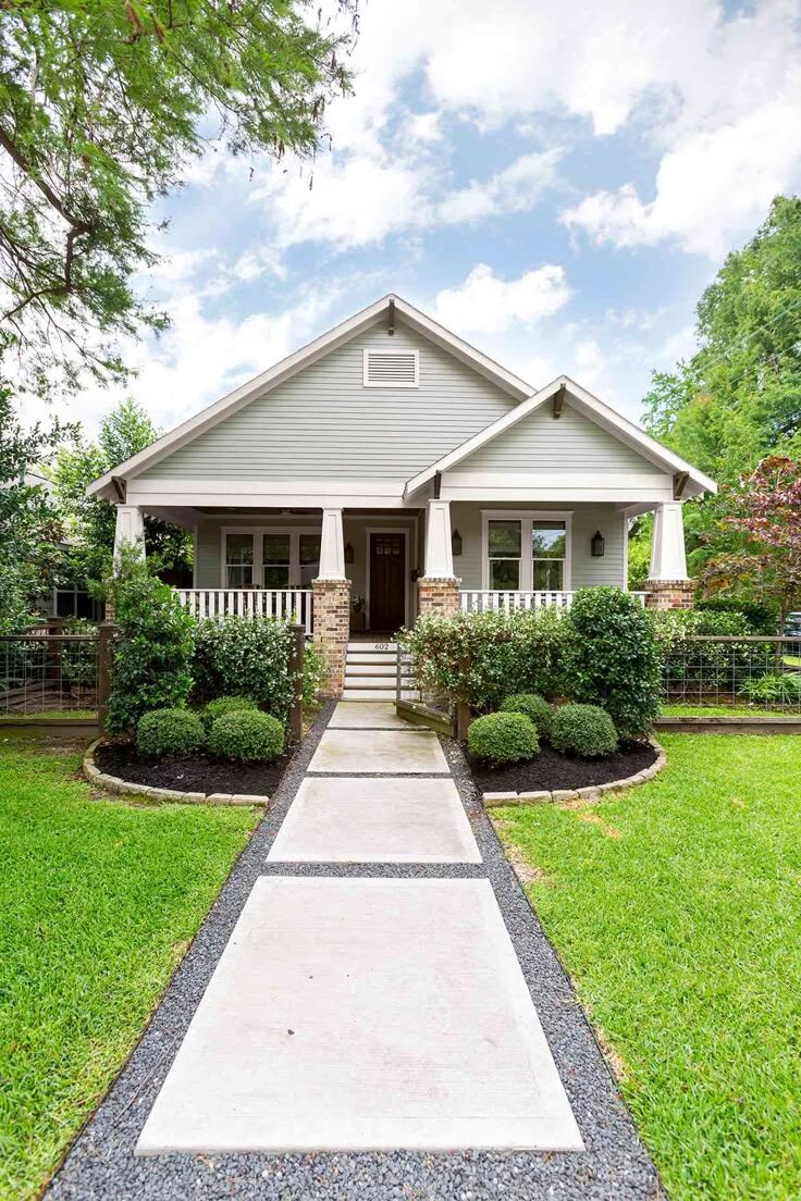 Charming bungalow-style home with light gray siding, brick and stone porch columns, manicured shrubs, and a modern concrete walkway leading to the entry.