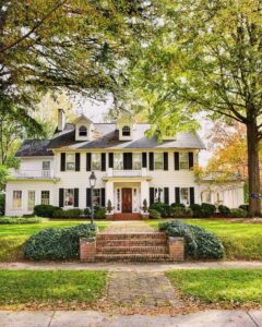 Colonial-style home with white siding, black shutters, symmetrical windows, red front door, and a brick walkway surrounded by manicured landscaping and mature trees.