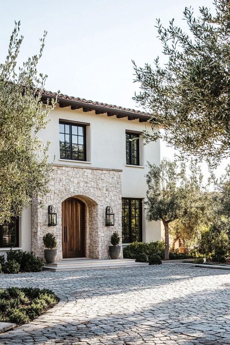 Mediterranean-style home with light stucco exterior, arched stone veneer entryway, wood front door, black framed windows, and cobblestone driveway.