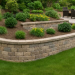 Curved, multi-tiered stone retaining wall in a landscaped backyard, featuring neatly trimmed shrubs, ornamental grasses, and a paved patio seating area.