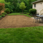 Cleared backyard area prepared for patio installation with green lawn, garden beds, and wooden fence on a sunny day.
